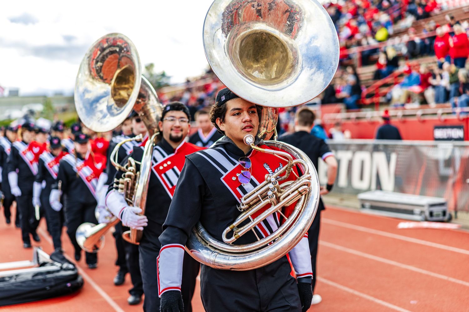 Marching band members perform with brass instruments on a track field during a halftime show at a stadium.