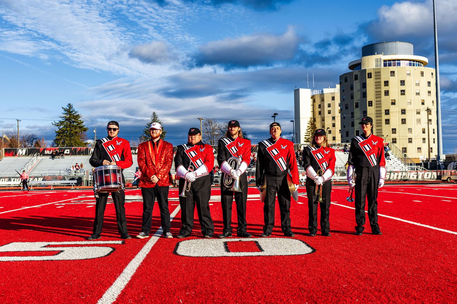 A marching band in red uniforms performs a field show formation on a bright red artificial turf field.