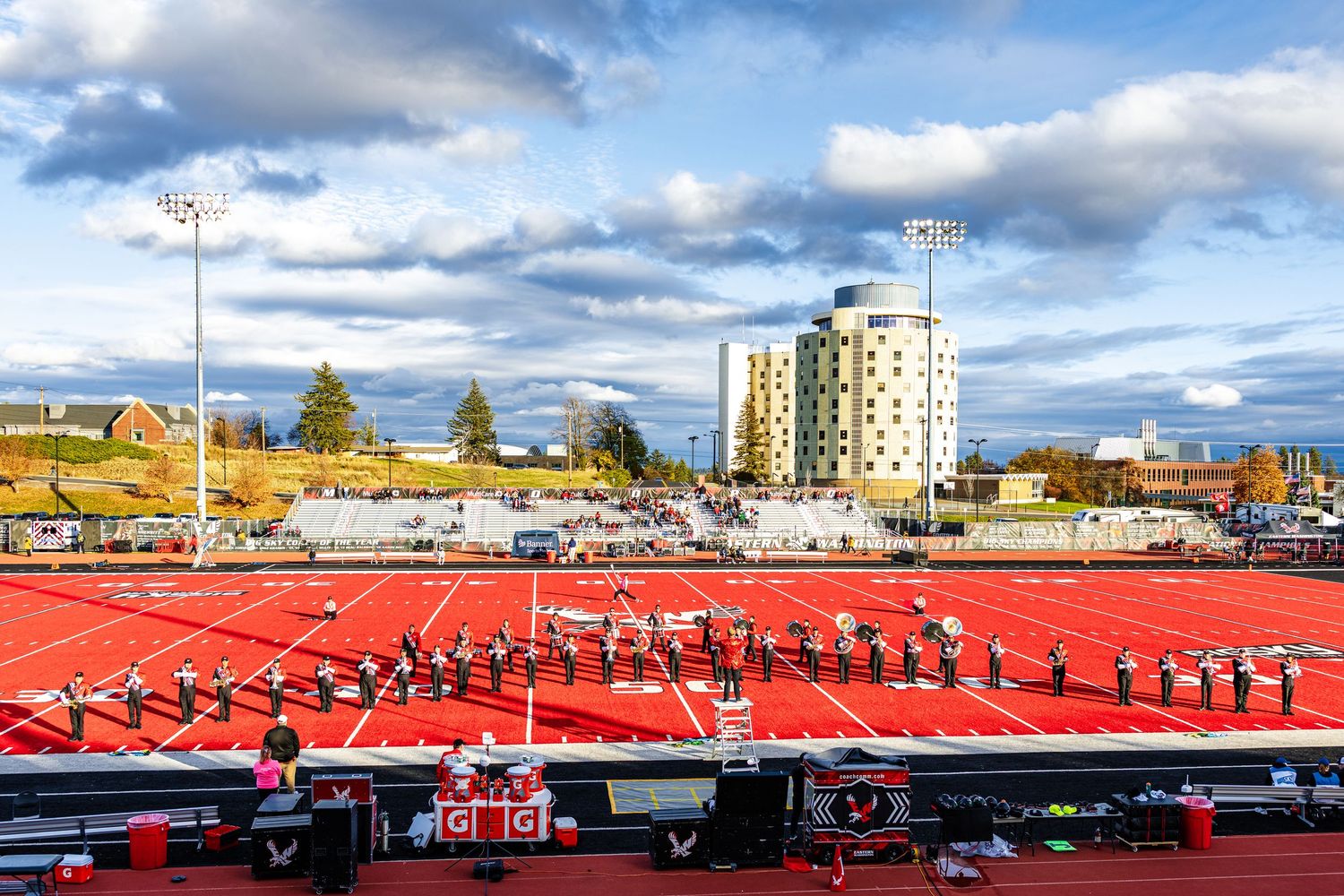 A bright red stadium with rows of empty seats overlooks a cityscape with a tall building under dramatic cloudy skies.