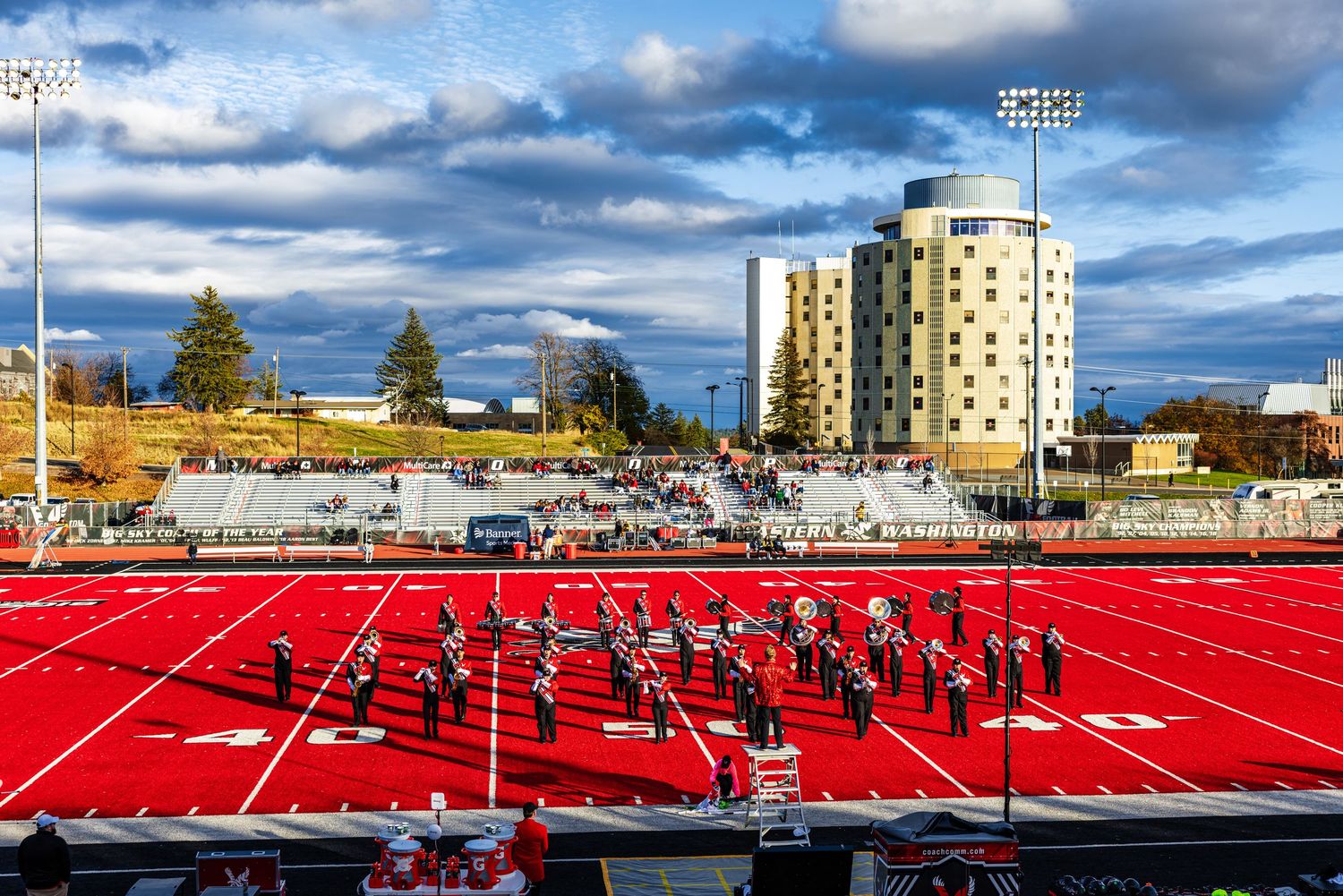A bright red running track and sports field with stadium seating and buildings visible under a cloudy blue sky.