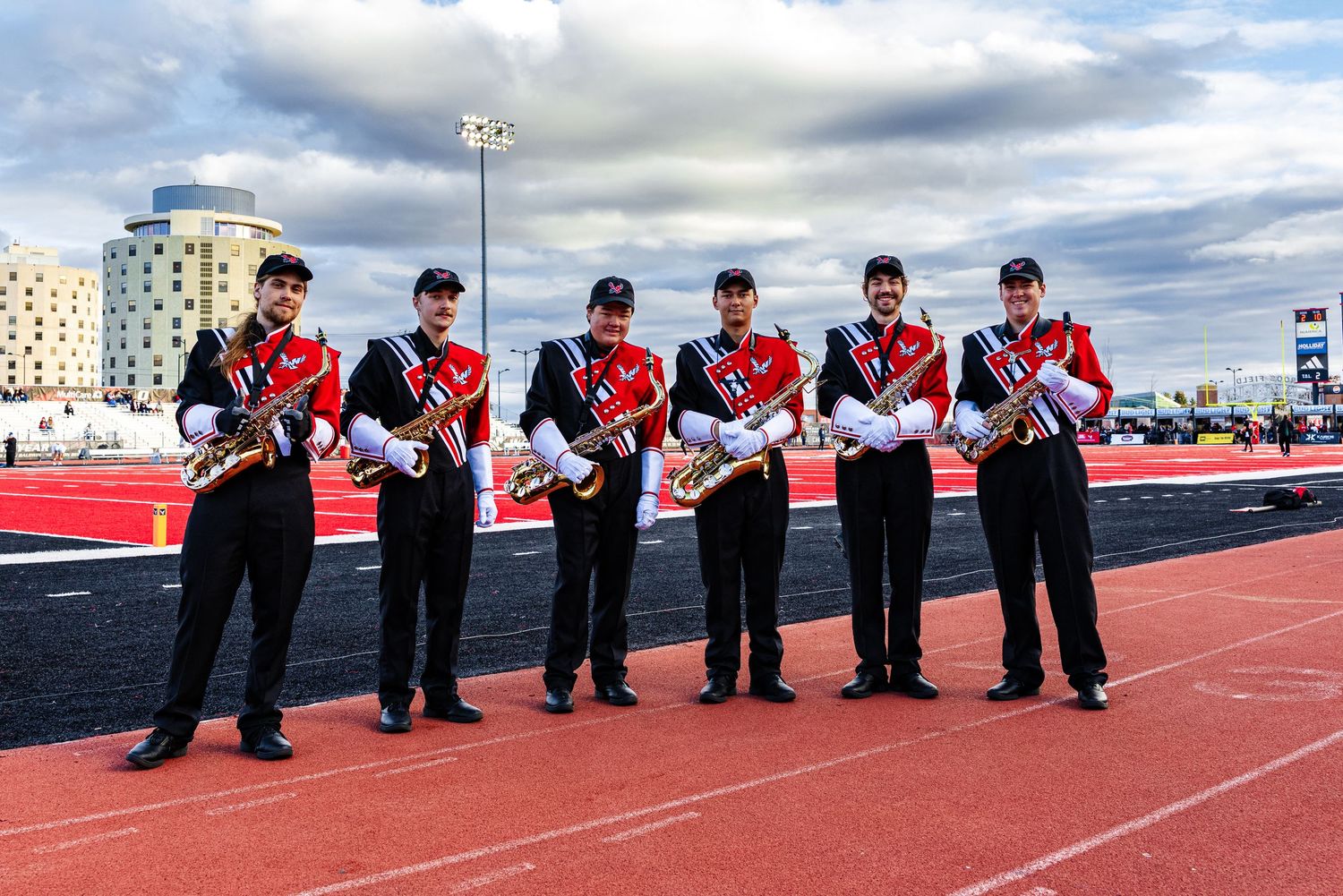 A line of uniformed marching band members in red and black attire stand with their instruments on a red track field.