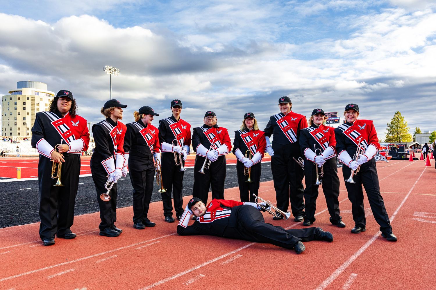 Marching band members in red and black uniforms pose together on a running track under blue sky with clouds.