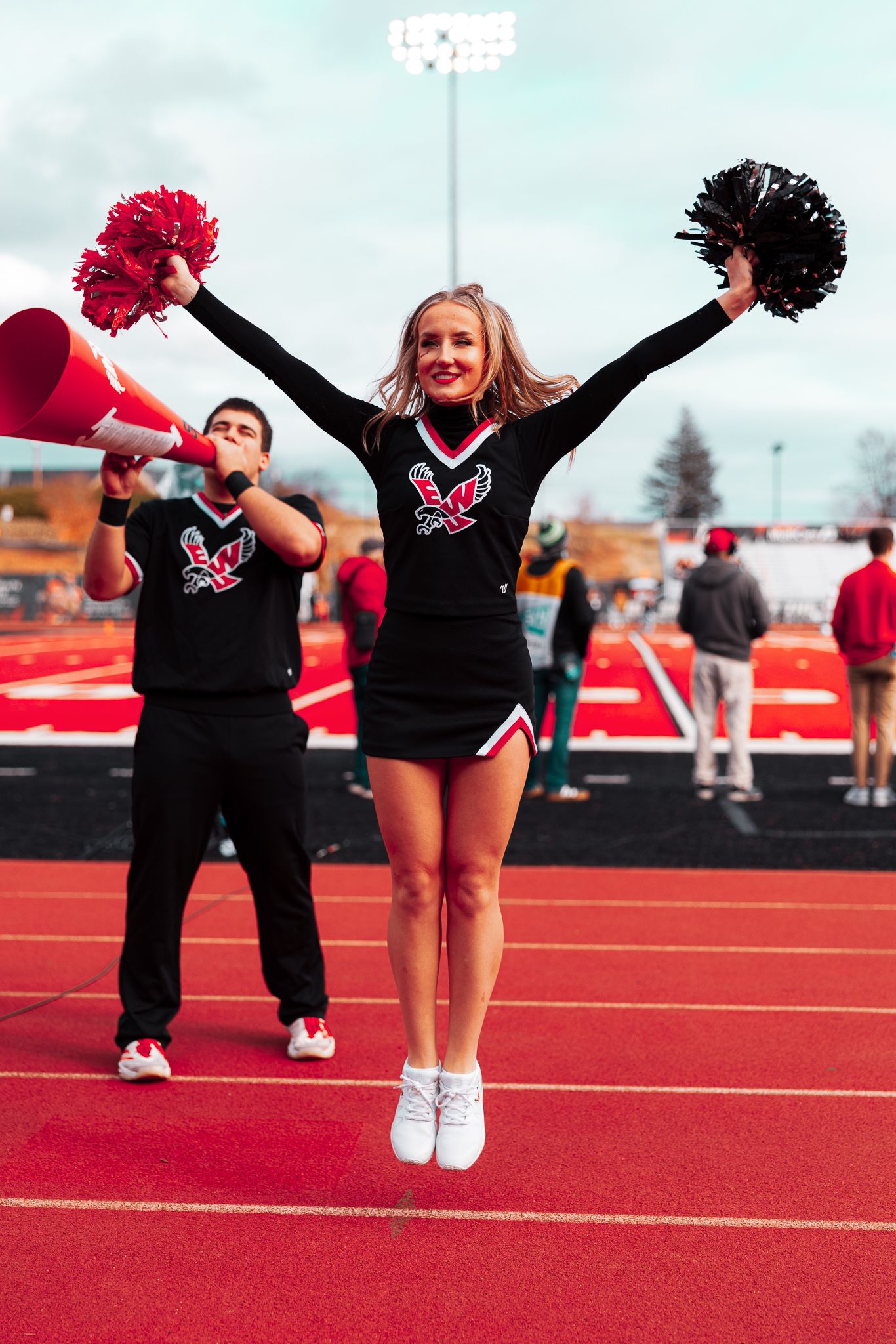 Cheerleaders in black uniforms perform high-energy jumps and poses on a bright red athletic field surface.