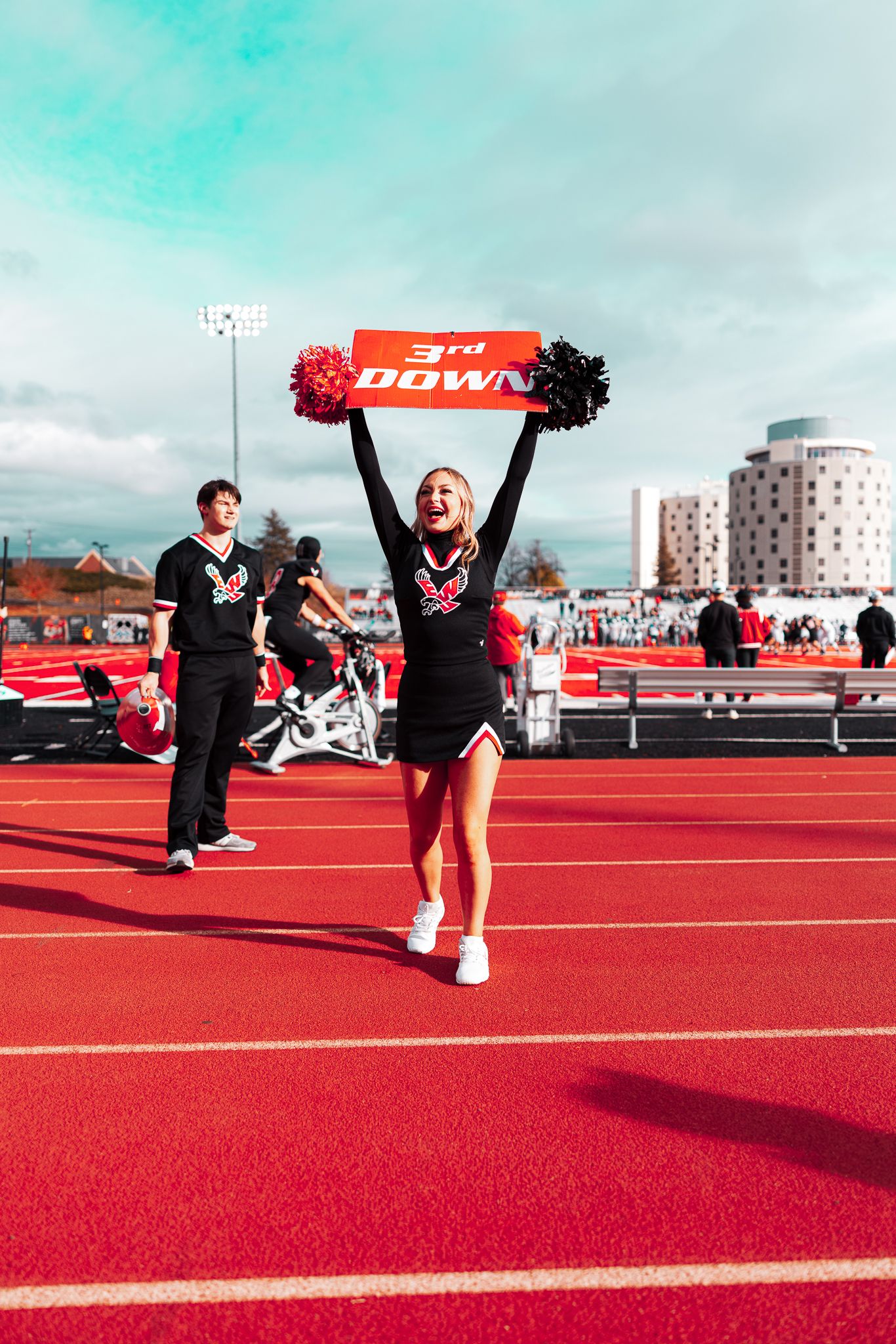 Cheerleaders perform synchronized jumps and movements on a bright red athletic track under a cloudy sky.