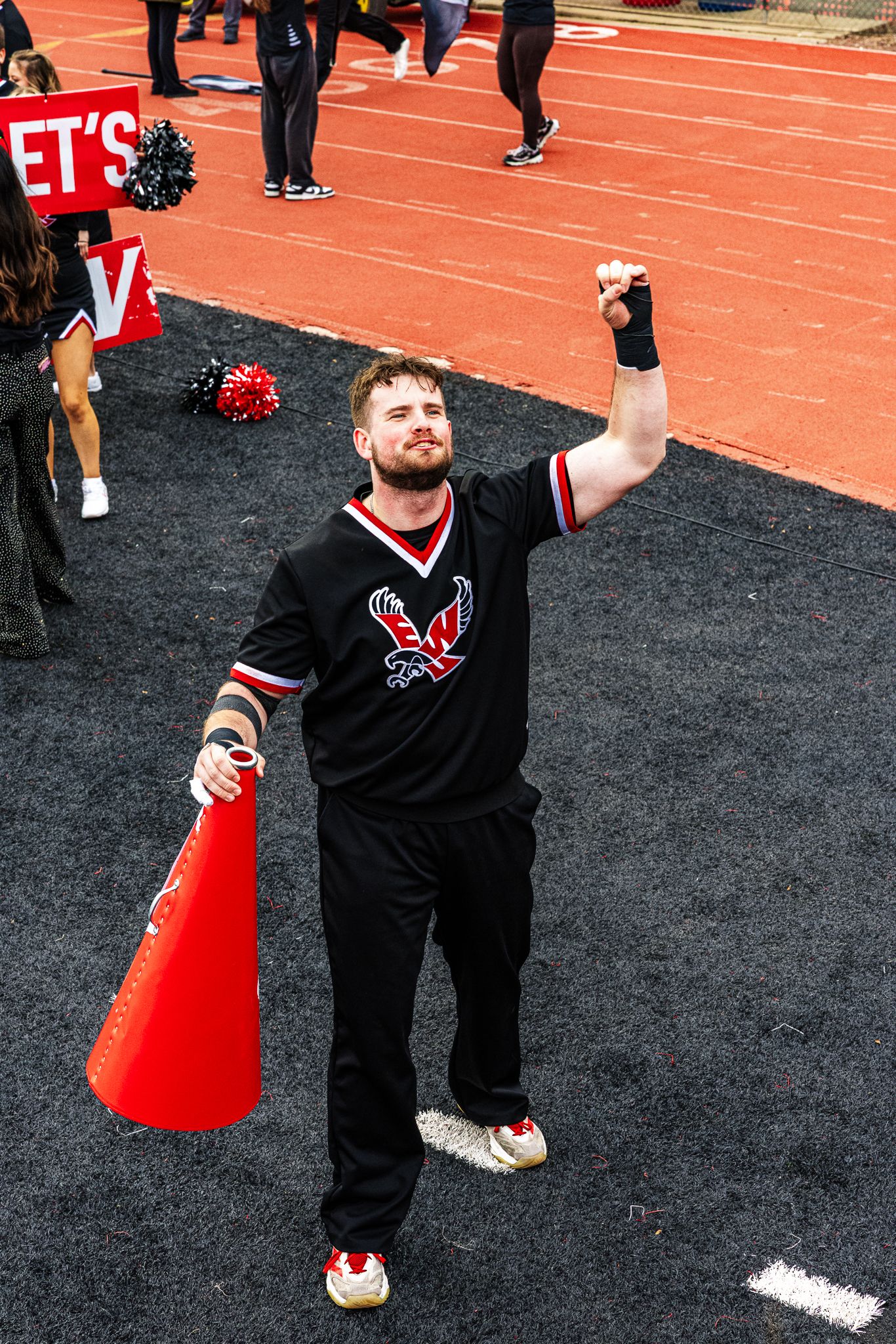 Cheerleader in black uniform with red megaphone performs routine on track field sideline.