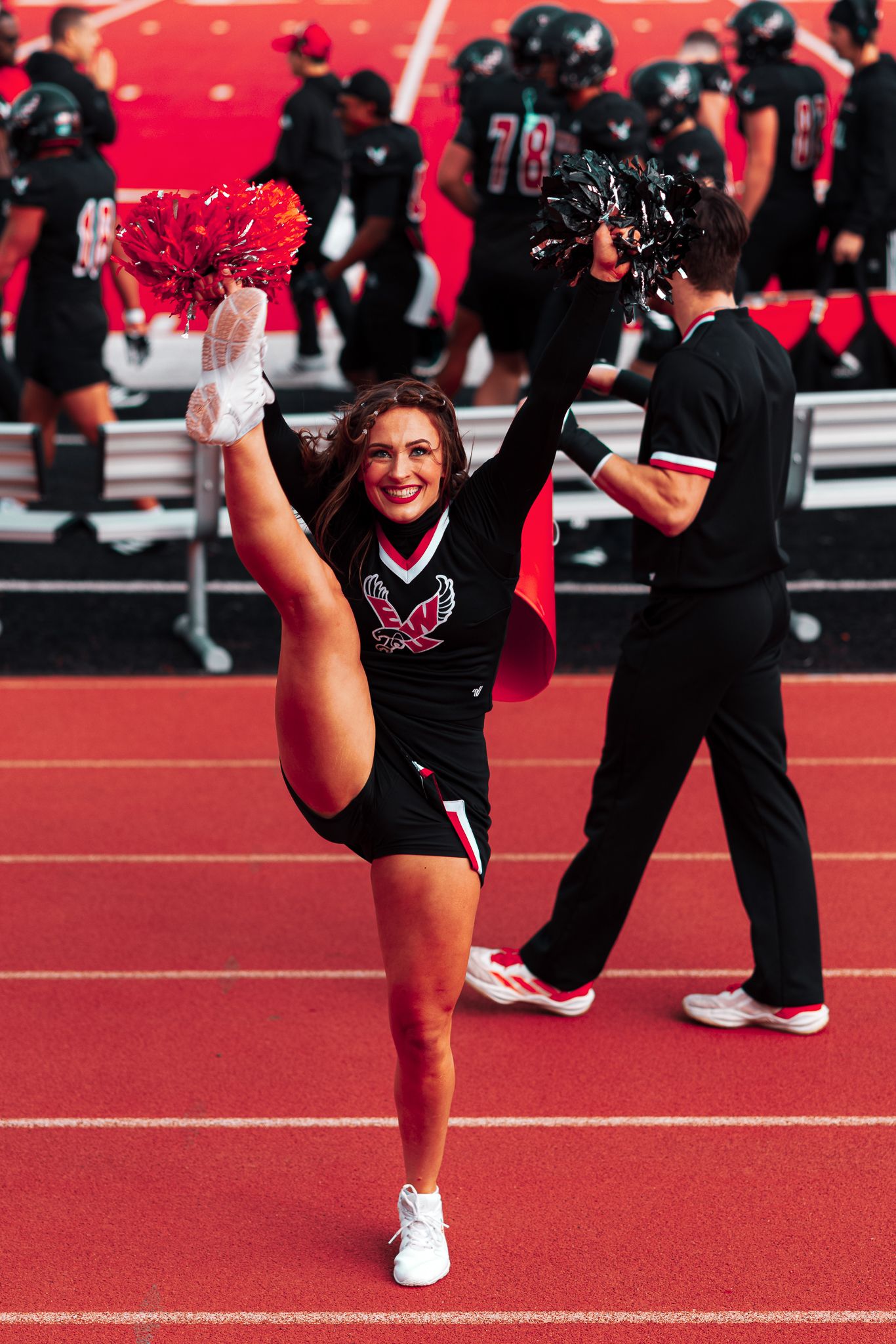 Cheerleader in black uniform performs high kick routine on red track field during sporting event.