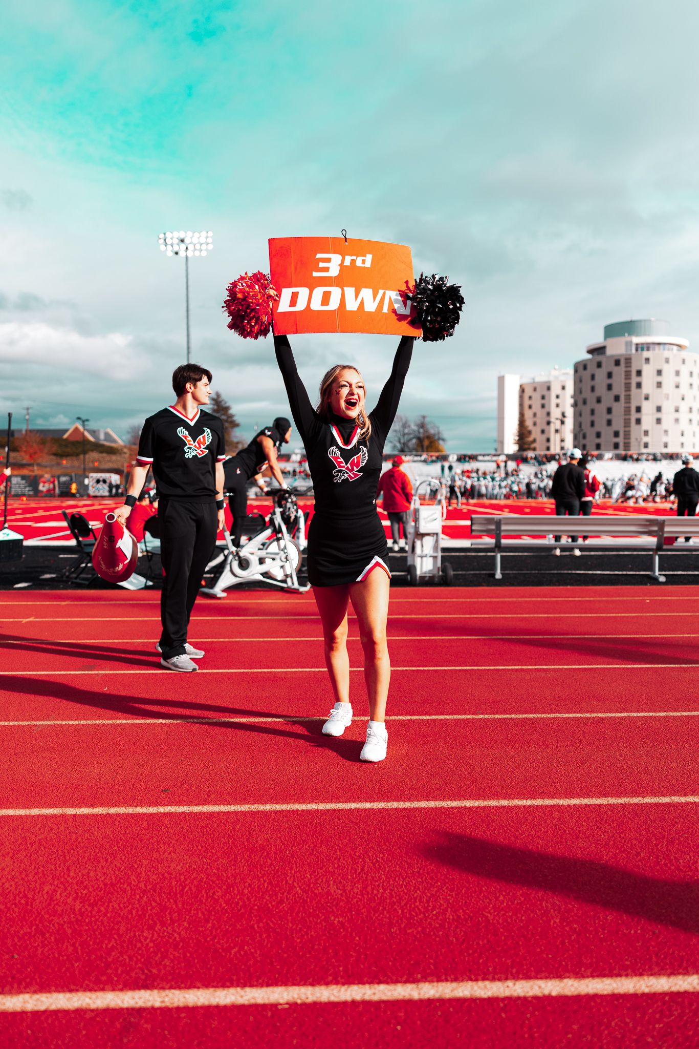 Cheerleader celebrates with red and black pom-poms and 3rd down sign on bright red athletic track during game day.