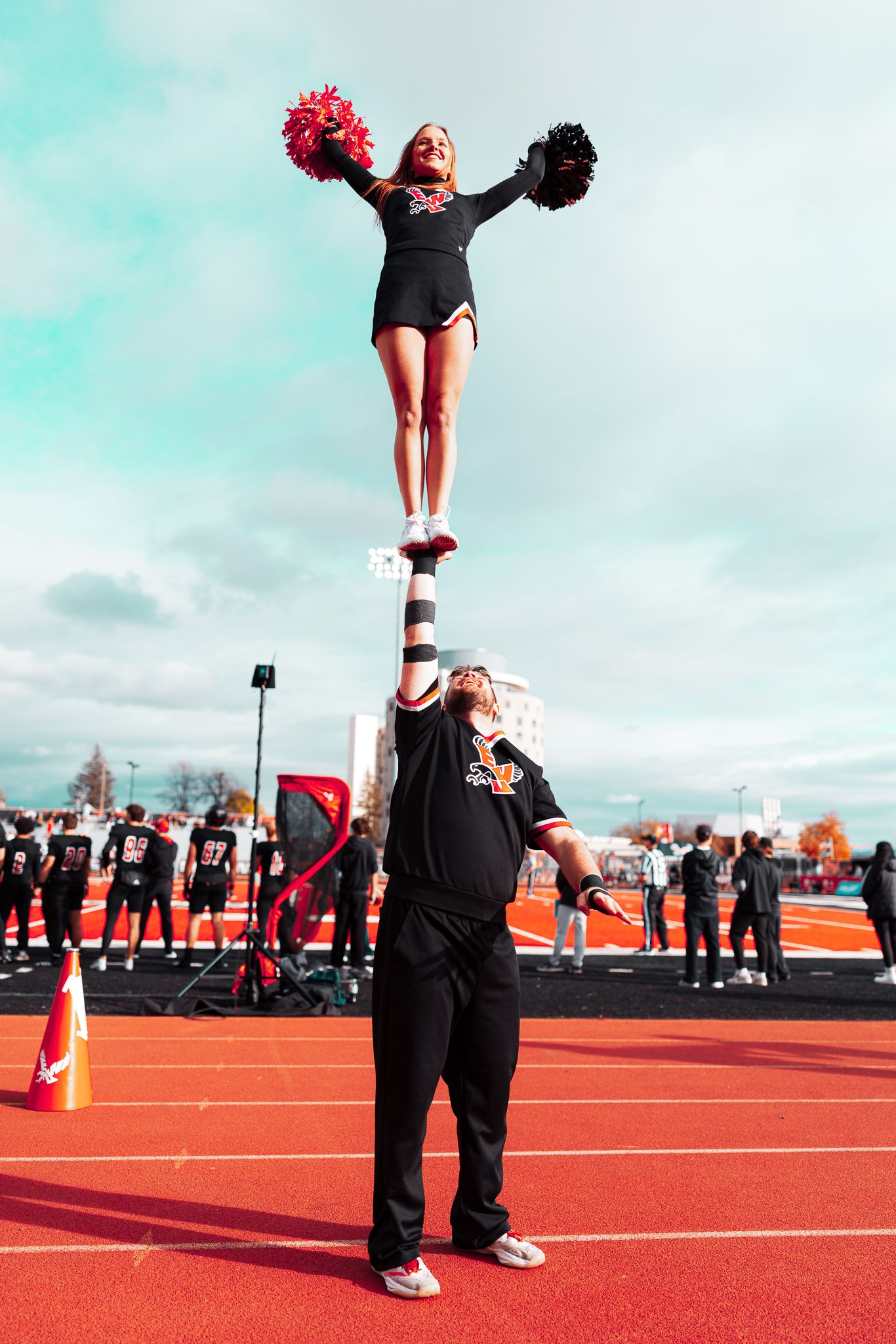 Cheerleaders perform a stunt sequence on a bright red athletic field showing progressive lifting movements.