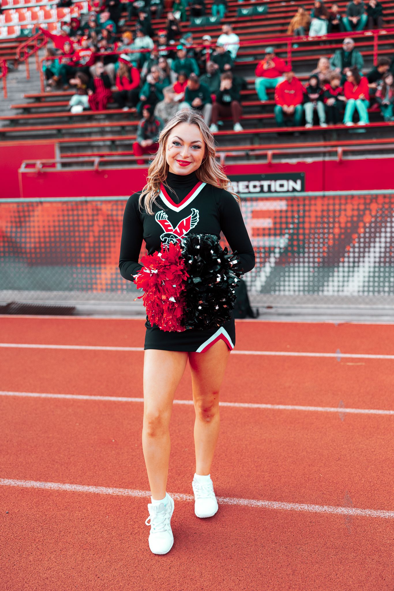 Cheerleader in black uniform with red pom poms performs on red athletic track during sporting event.