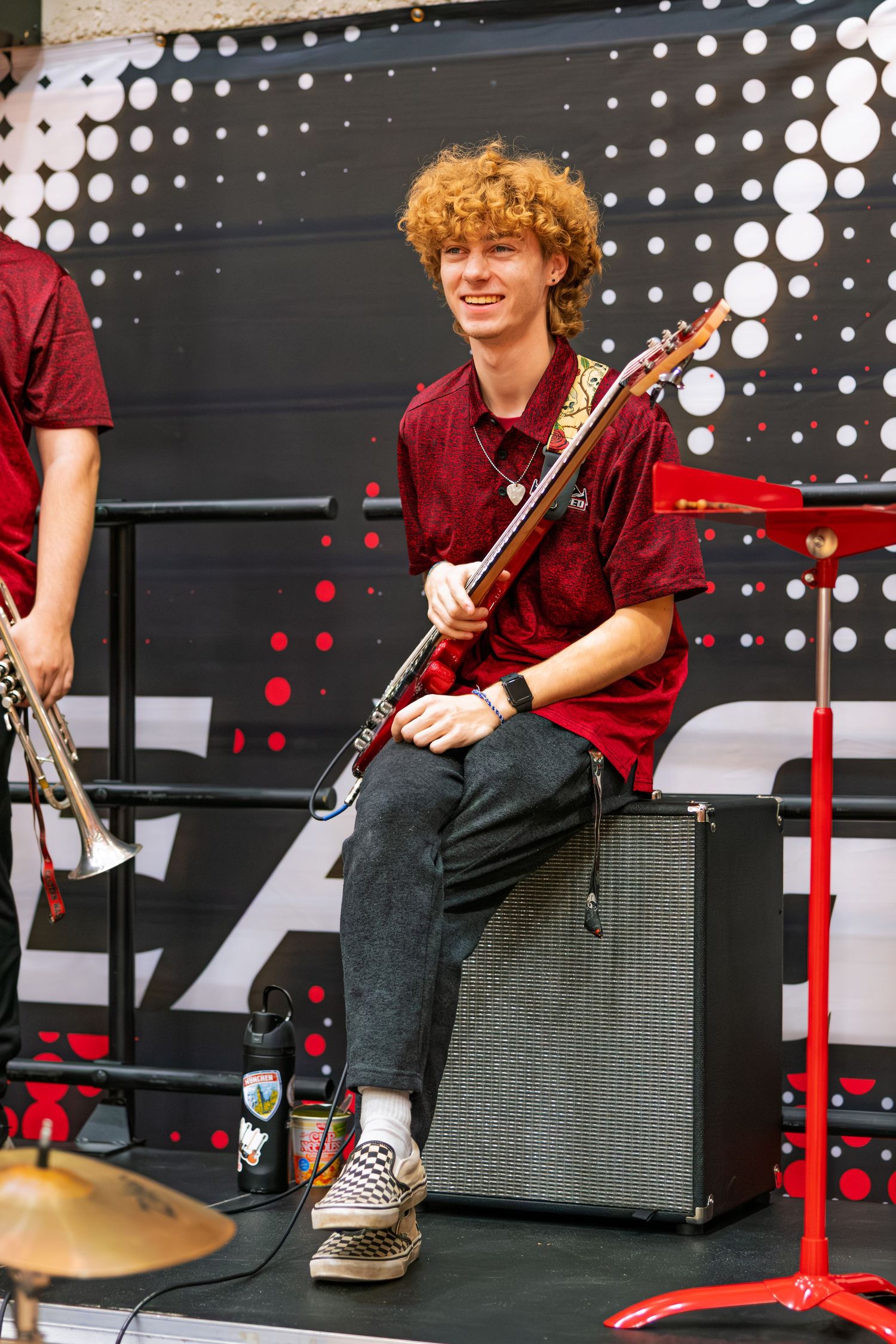 Musicians in burgundy shirts perform on stage with instruments and equipment against a dark background with illuminated dots.