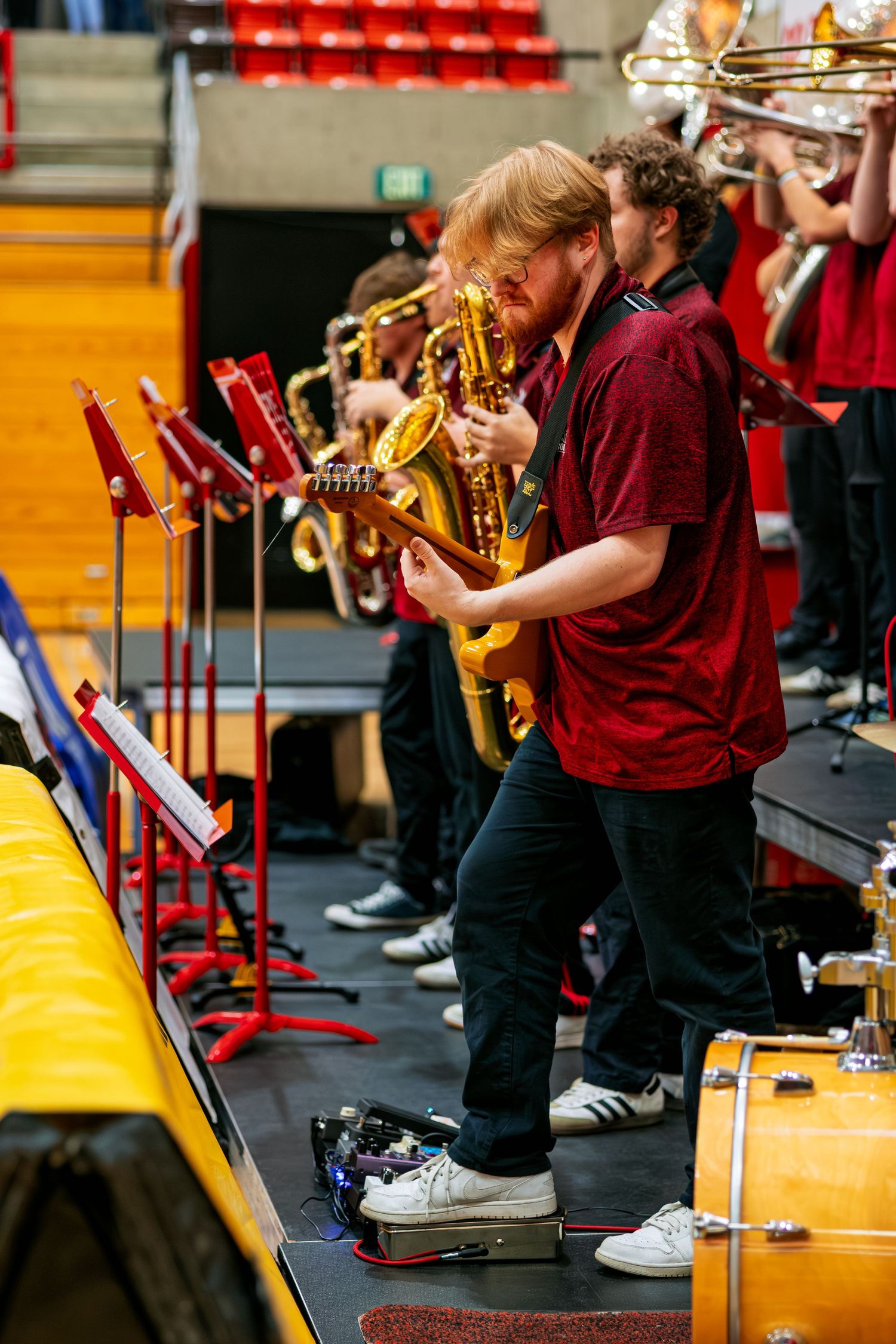 Students in maroon uniforms play brass instruments while seated in a row during a band performance.