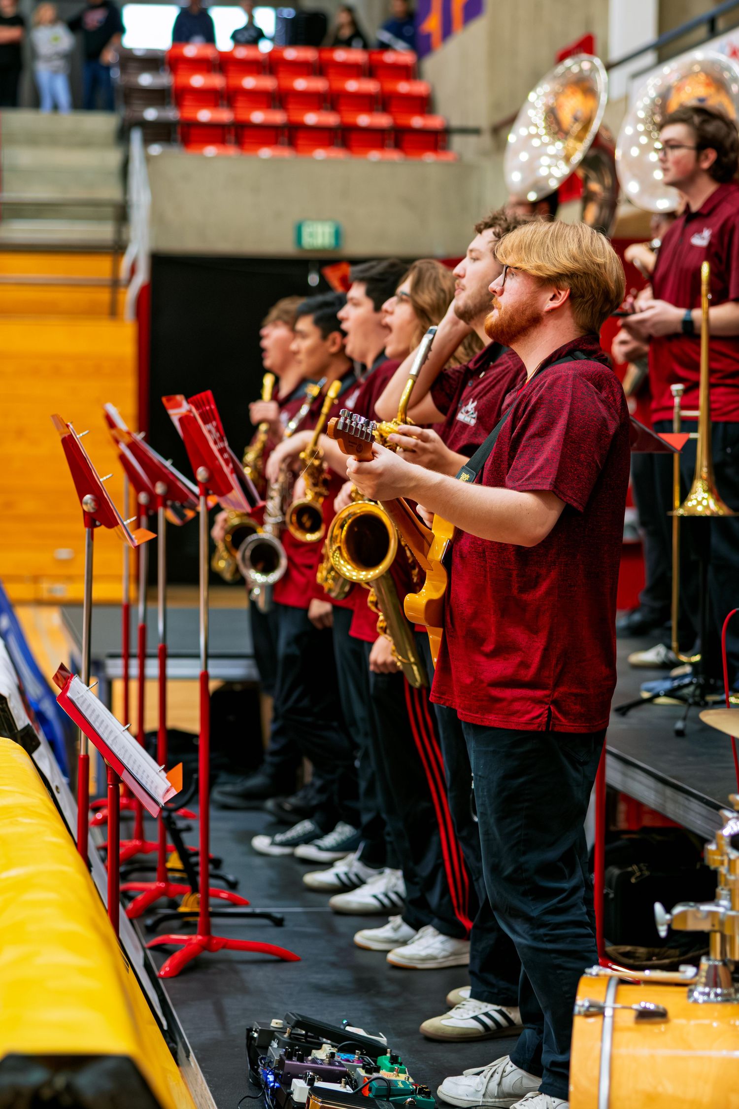 College pep band musicians in maroon uniforms perform with saxophones and brass instruments during a basketball game.