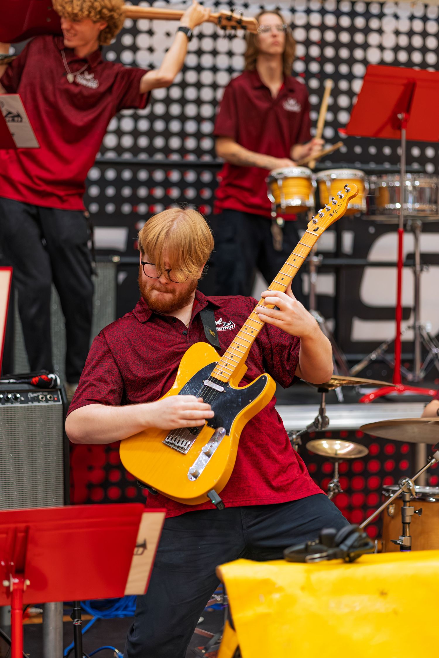 Musician in burgundy polo shirt plays yellow Telecaster guitar during live performance with band on stage.