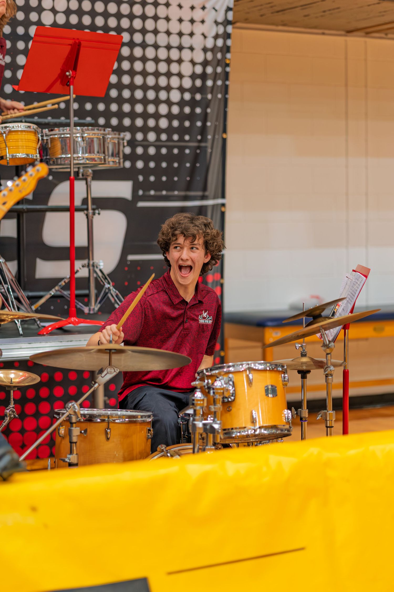 Drummer in a burgundy shirt performs energetically on a yellow stage with a dotted background pattern.