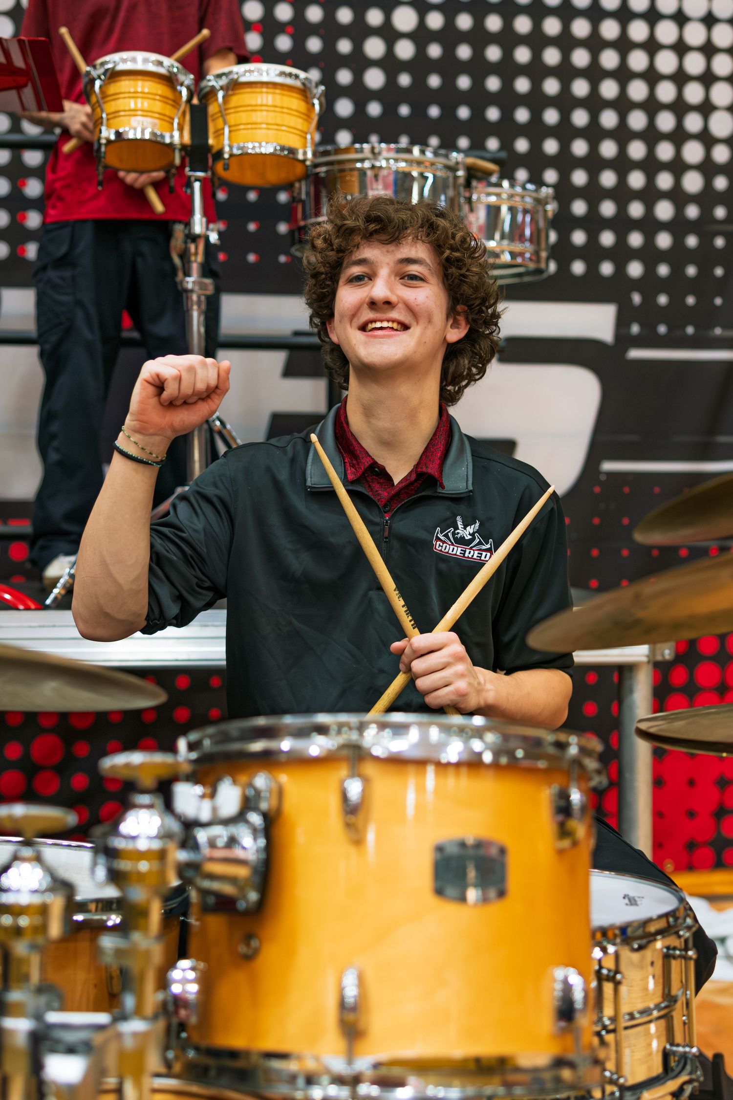 Drummer performs energetically behind a yellow drum kit on stage with spotlights and a dotted background pattern.