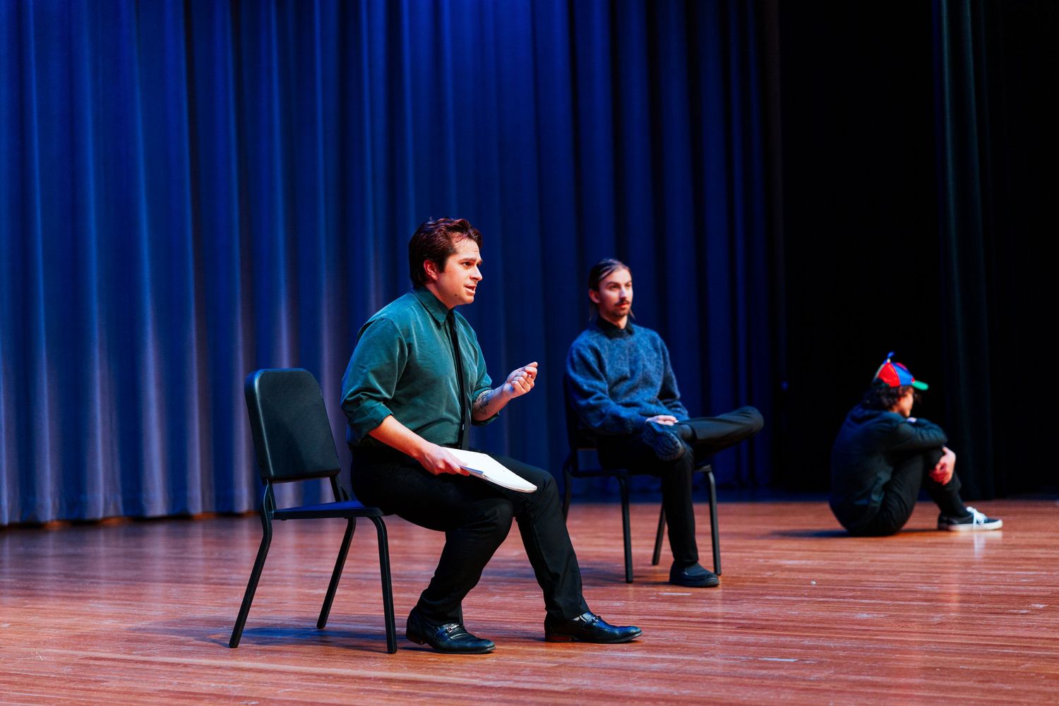 Three performers sit on a stage with blue curtains during a theatrical performance on a wooden floor.