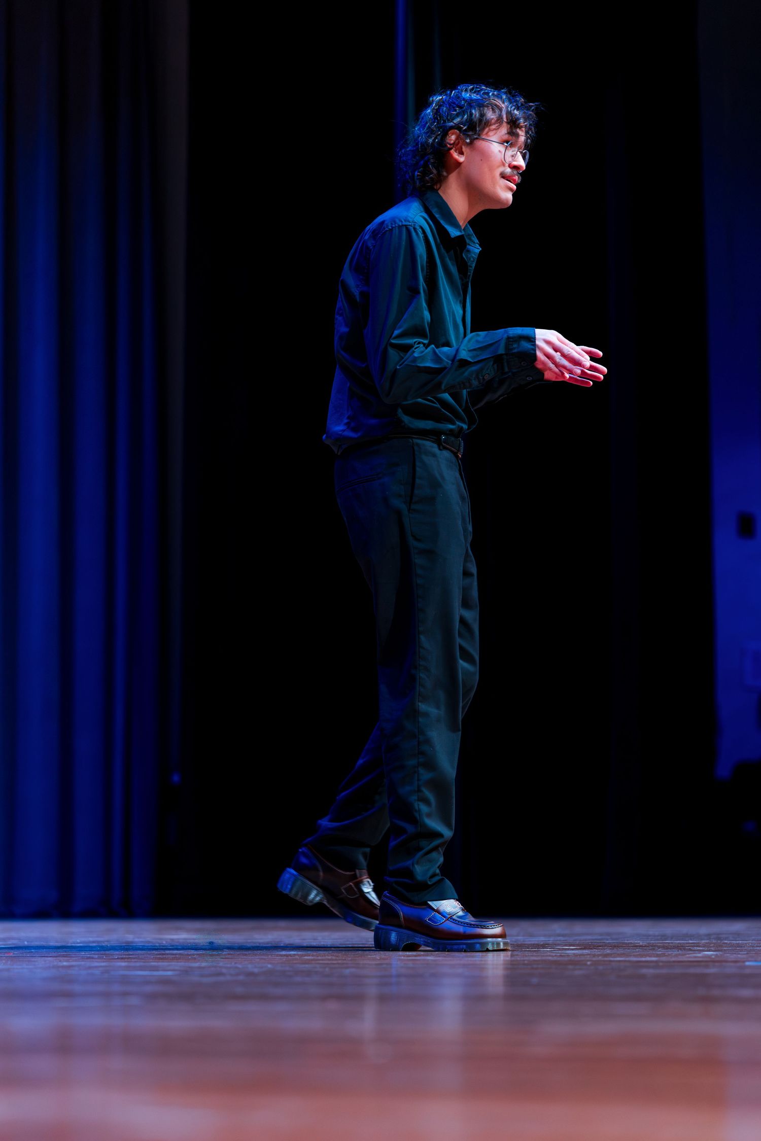 A performer in dark clothing stands on stage during a theatrical performance with dramatic blue lighting.