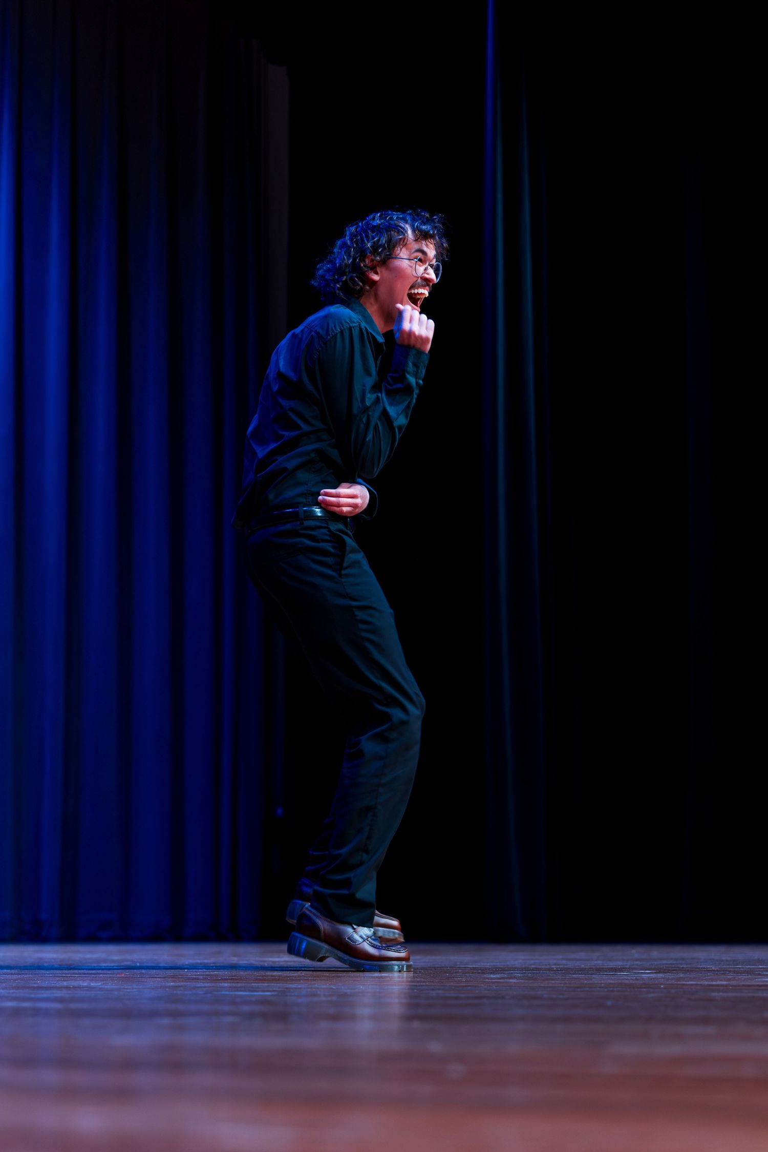 A dancer in black attire performs dynamic movements against a blue curtain backdrop on a wooden stage.