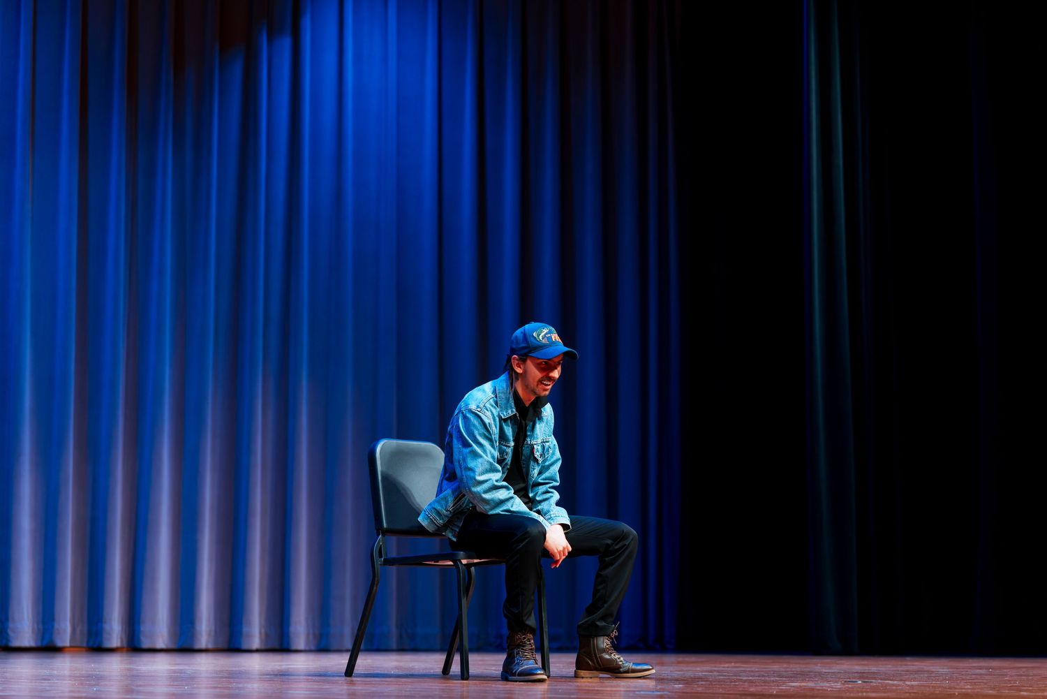 A performer in a blue shirt stands on stage in front of a dark curtain during a theatrical performance.