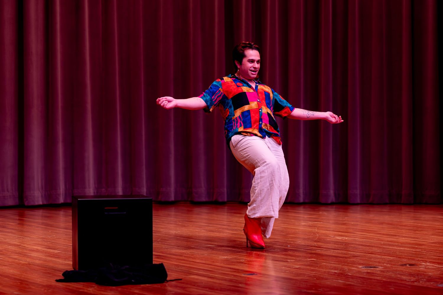 Performer in colorful costume dances energetically on wooden stage with red curtain backdrop.