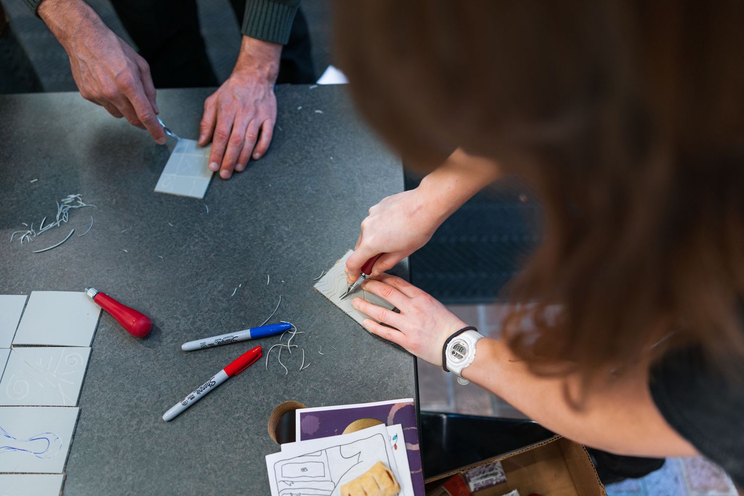 People working on a project with colored markers and papers spread out on a dark table surface.