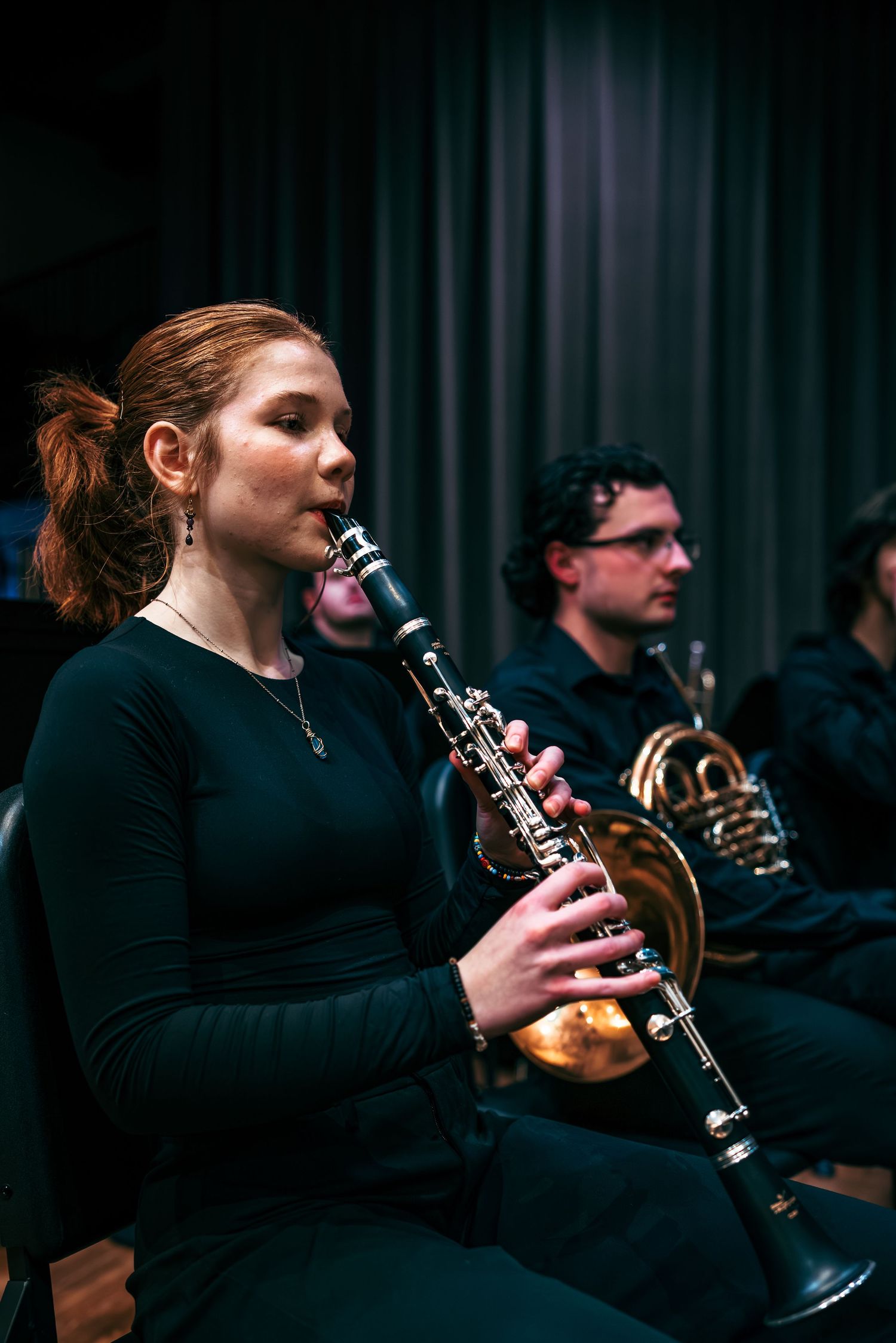 Musicians perform in an orchestra with clarinet and brass instruments during a darkly lit concert performance.