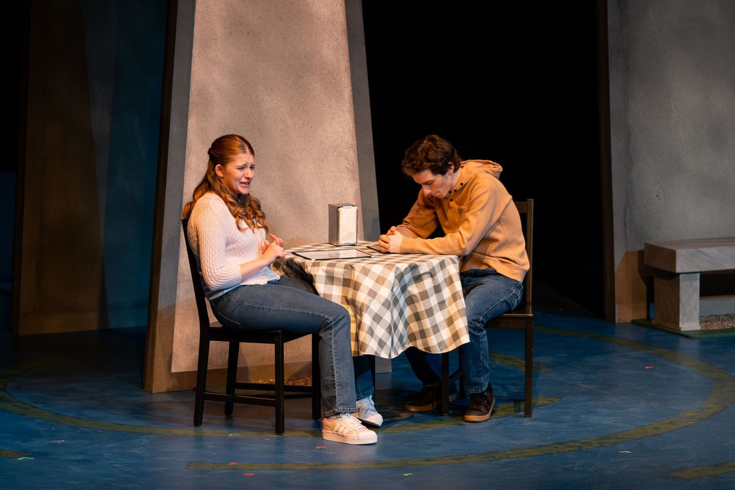 Two people sit on the floor engaged in intimate conversation during what appears to be a theater performance.