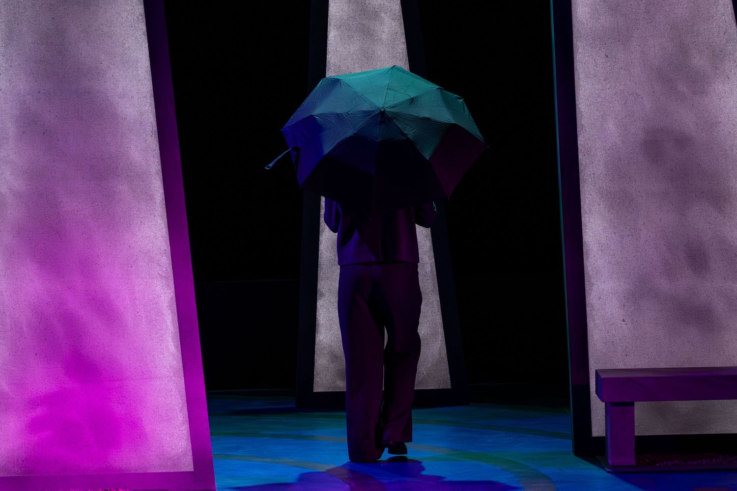A dark theater stage with dramatic purple and blue lighting illuminating vertical panels and silhouettes.