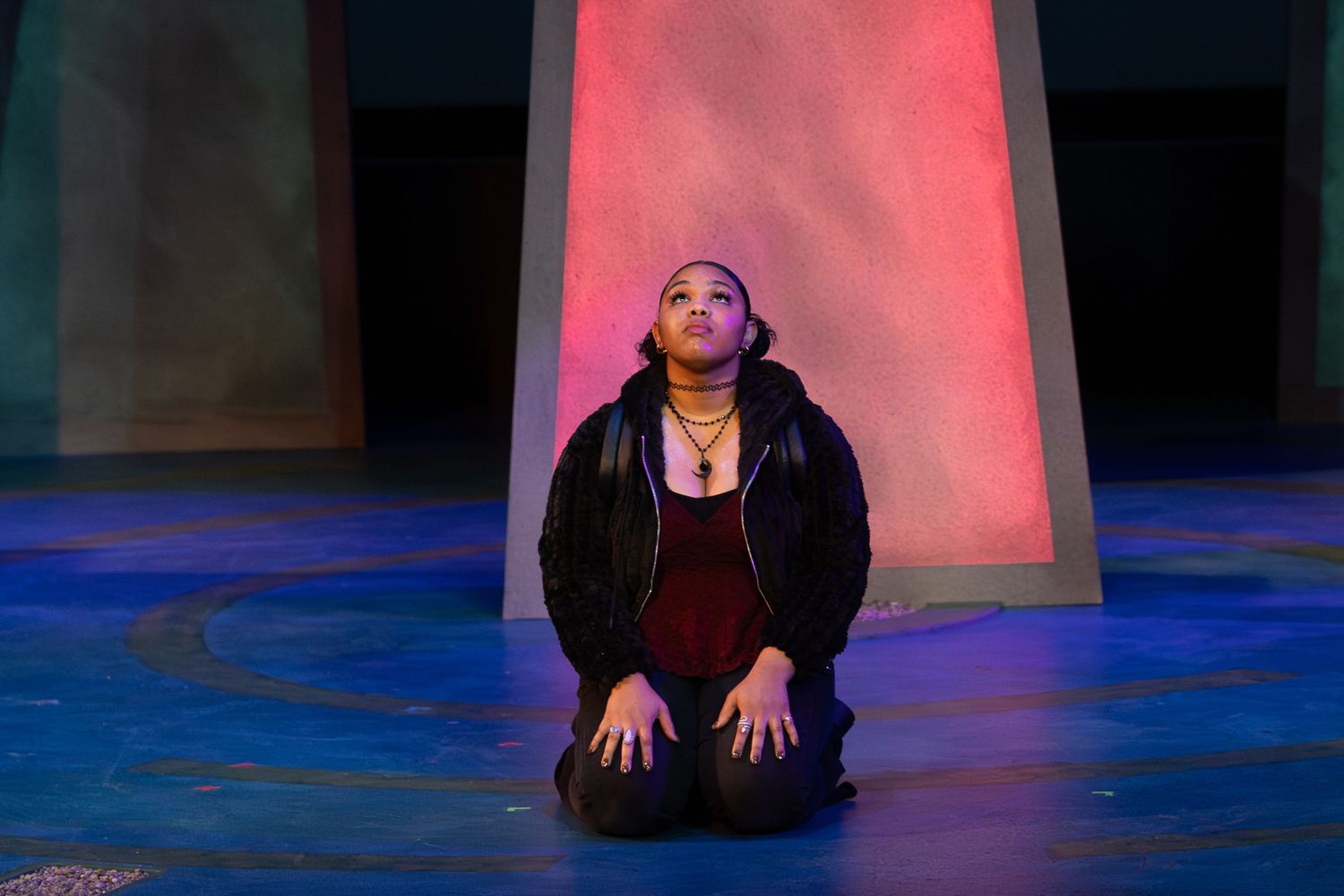A performer kneels on stage under dramatic pink and blue lighting during a theatrical performance.