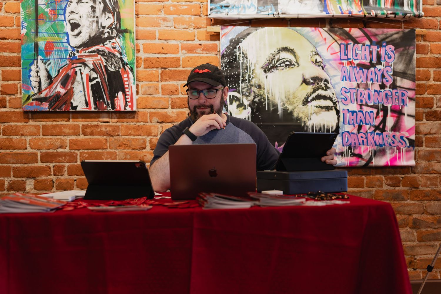 Man with beard and cap works on laptop at red table surrounded by colorful street art murals on brick wall.