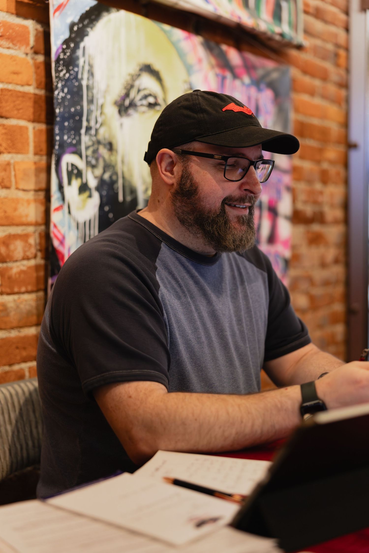 Bearded man wearing glasses and black cap works on tablet at rustic brick wall cafe with colorful street art mural.