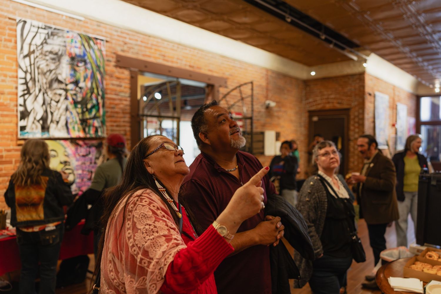 Two people admire artwork at an indoor gallery event with brick walls and paintings on display.