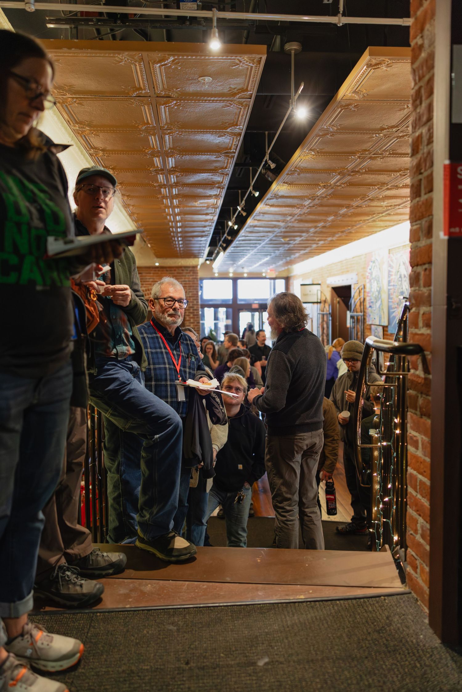 Crowd of people gathered in a rustic brick-walled venue with tin ceiling during a busy indoor event.