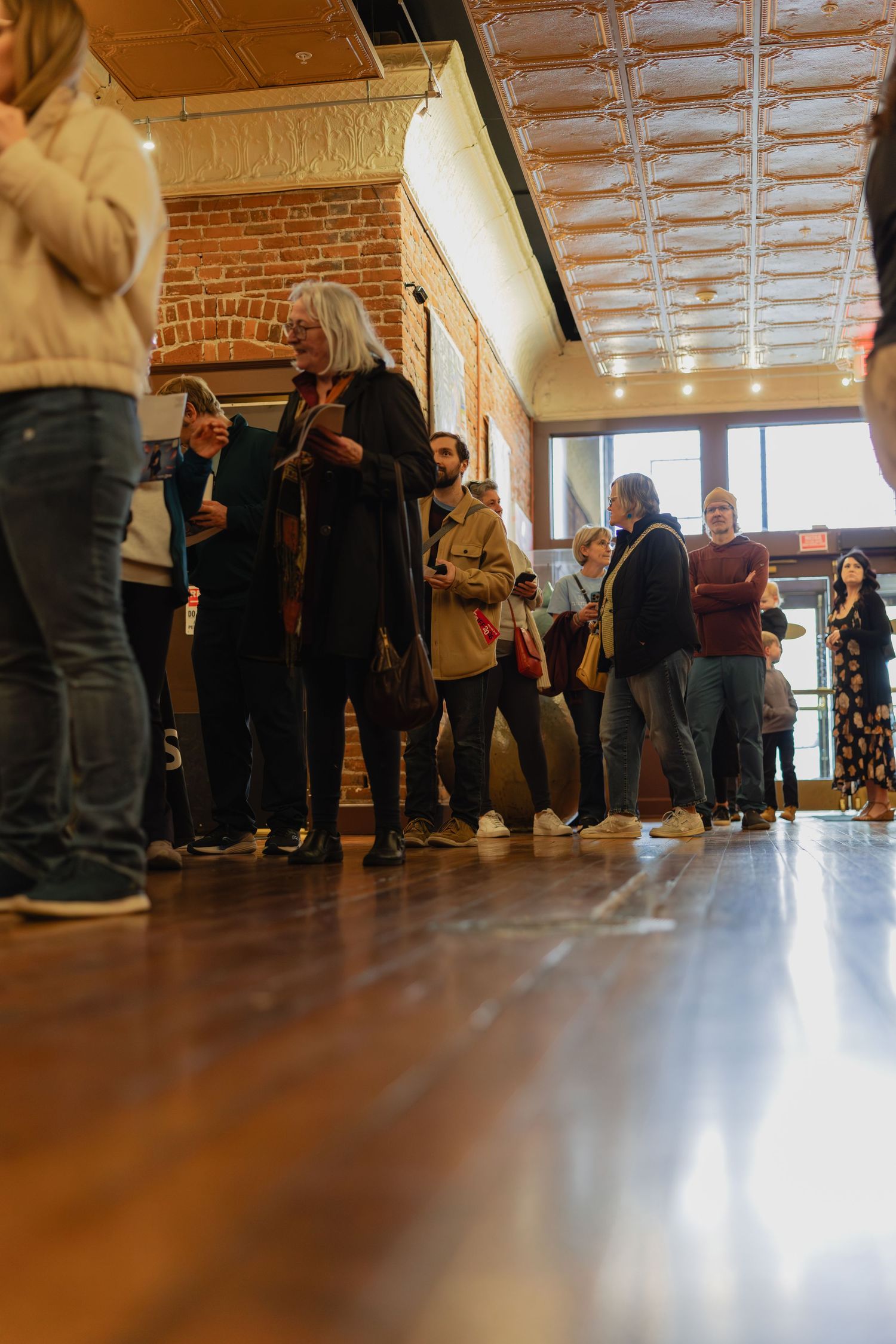 People standing in line inside a rustic venue with brick walls, tin ceiling, and hardwood floors.