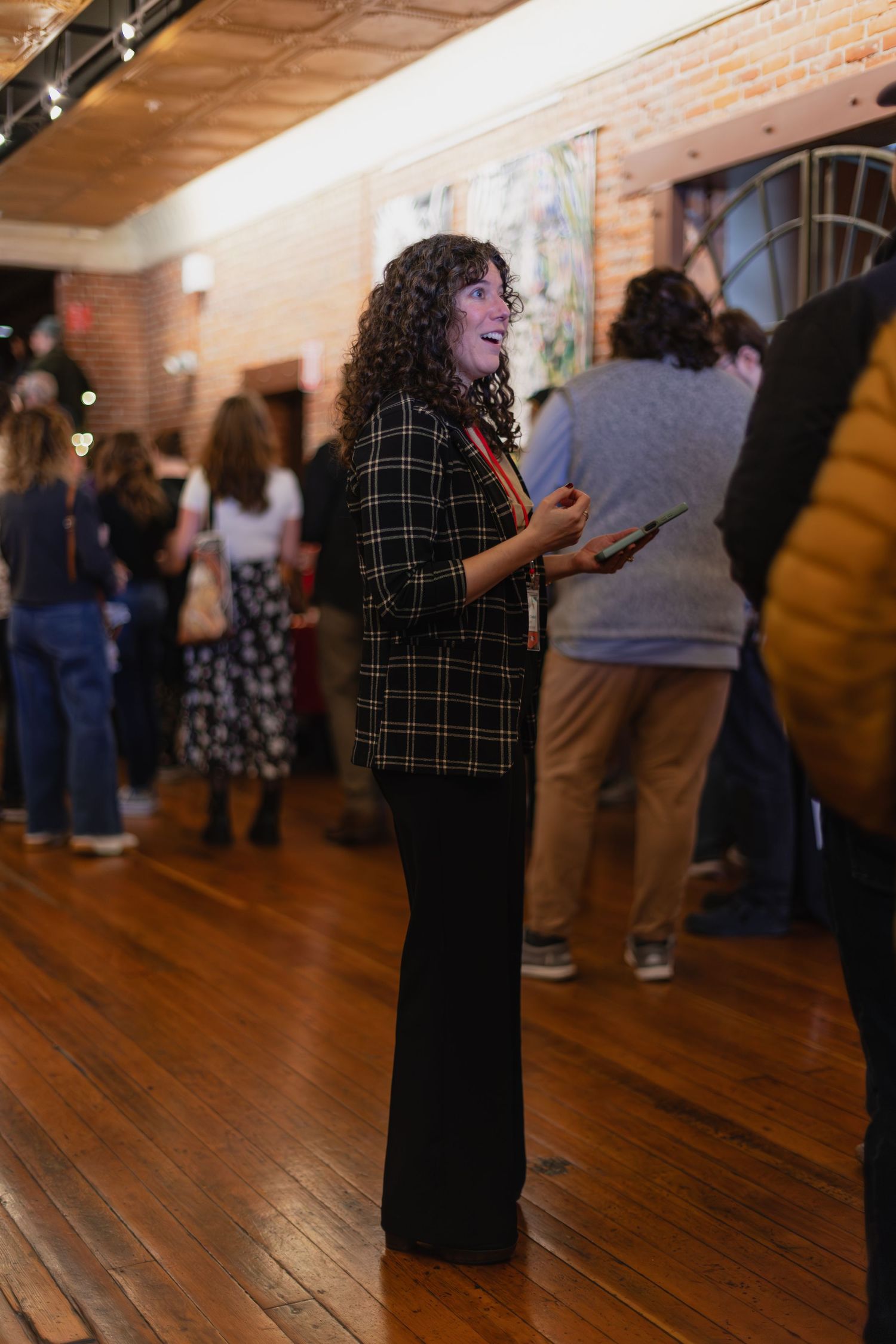 Woman with curly hair in plaid blazer and black pants smiling at an indoor art gallery event.