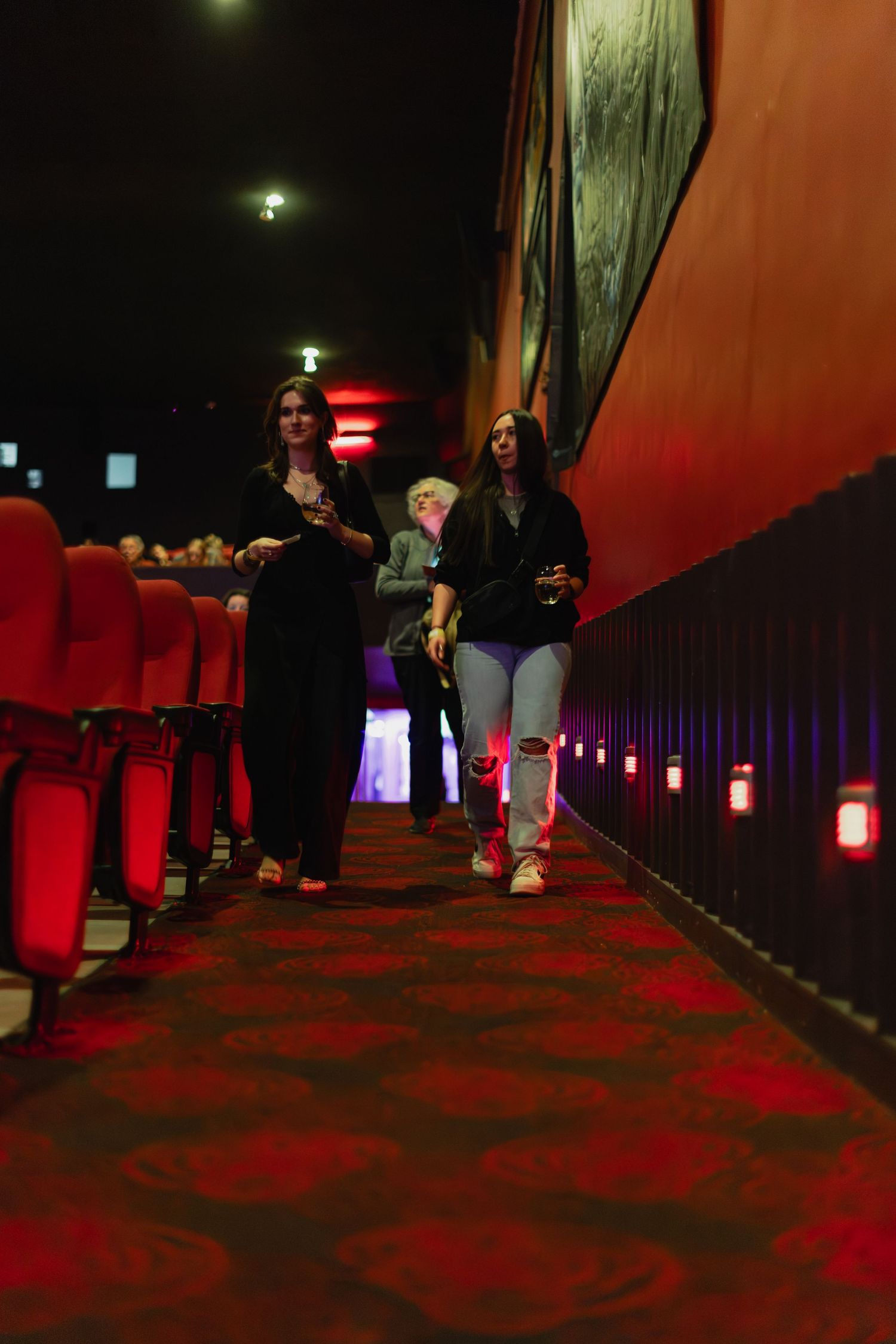 Two women walking down aisle of dark cinema theater with red seats and glowing floor lights.