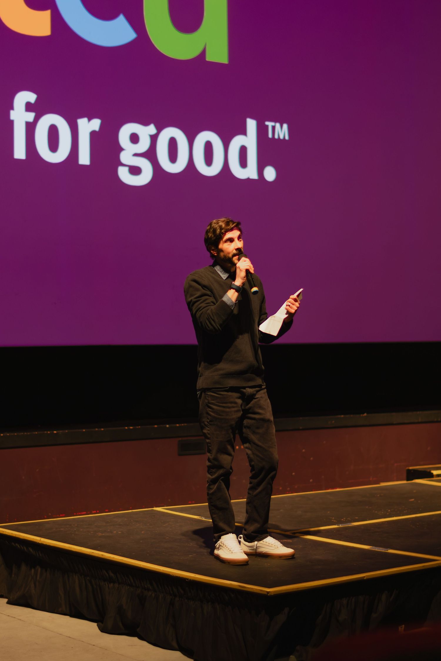 Man speaking on stage with microphone in front of purple screen displaying 'for good' slogan.