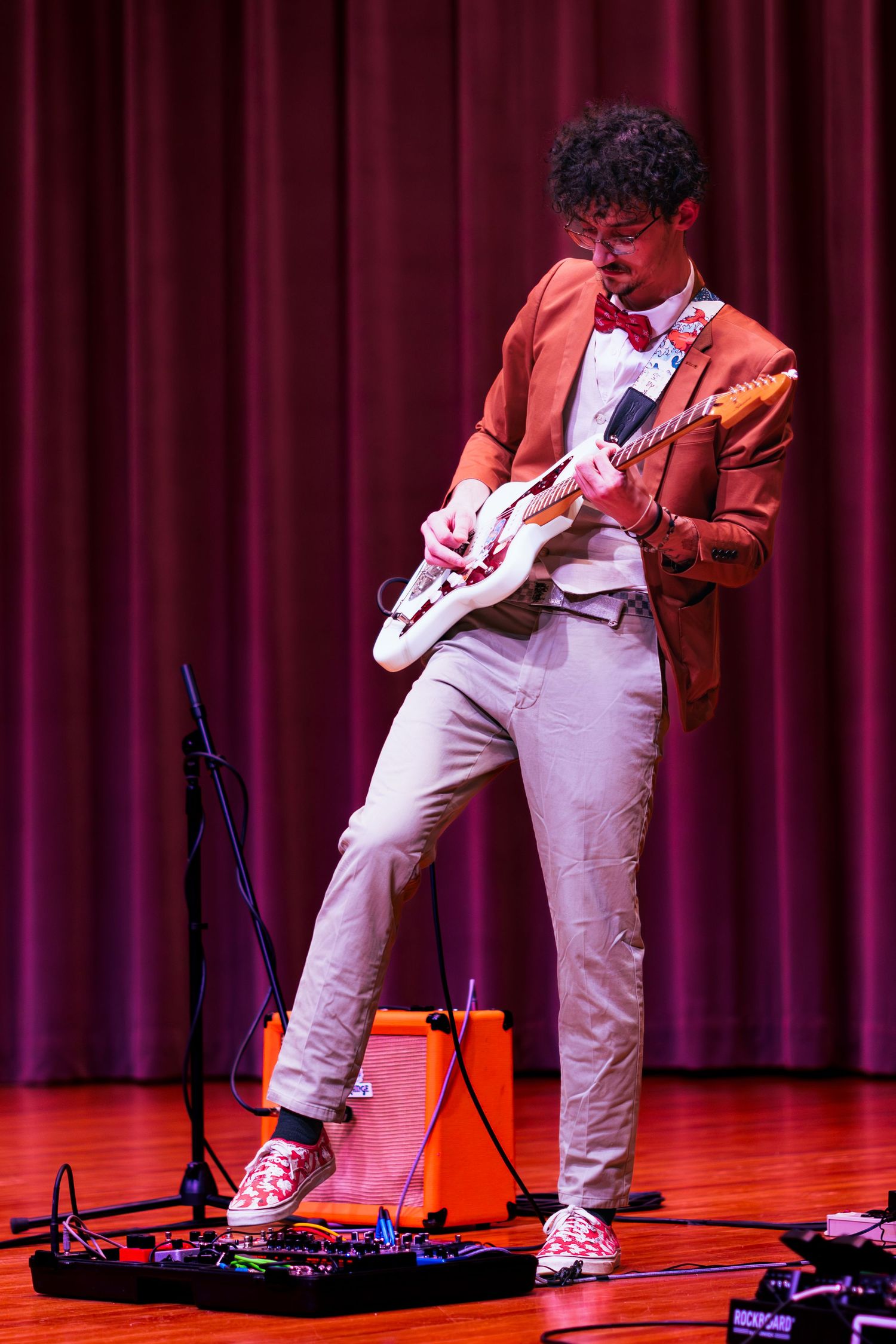 Musician in orange blazer and white pants plays electric guitar on stage with red curtain backdrop during performance.