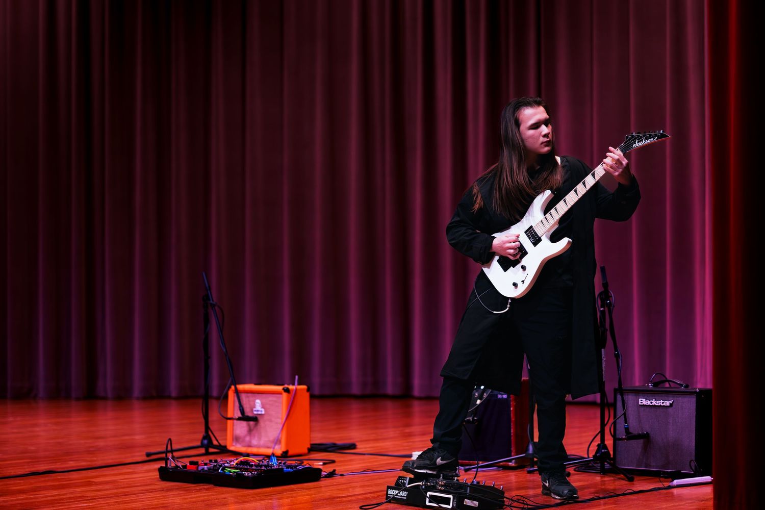 A guitarist performs on stage with dramatic red curtain backdrop and spotlighting during a live concert.