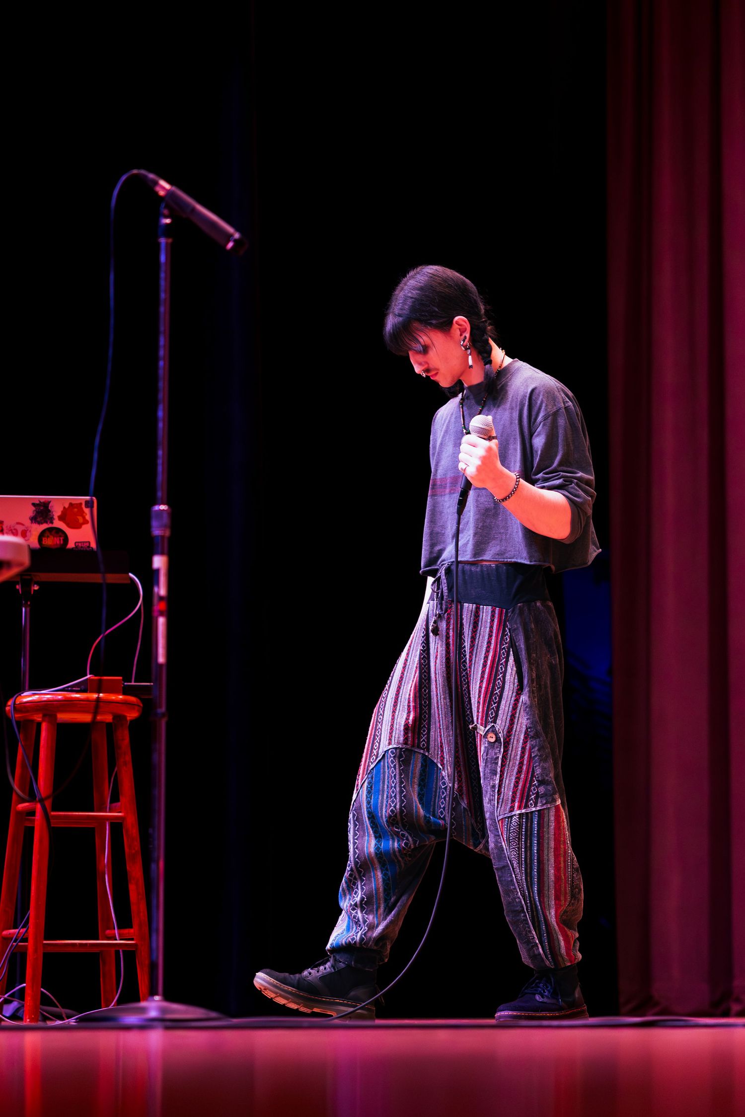 A performer in dark clothing stands on stage illuminated by dramatic red and blue lighting during a theatrical production.