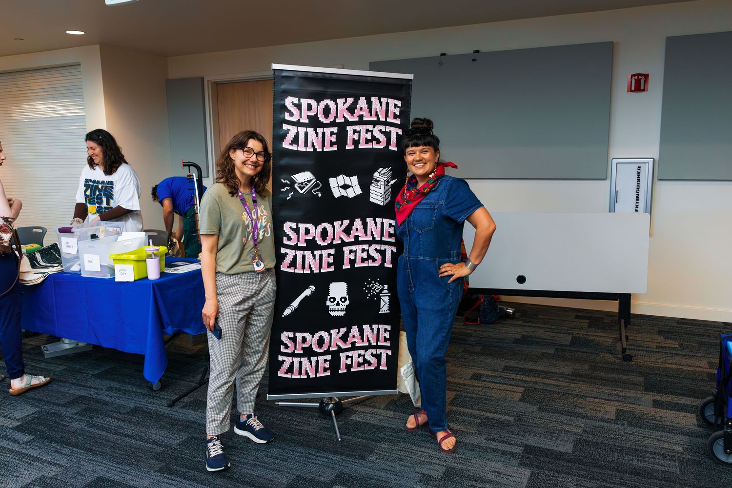 Two people stand next to a black banner for the Spokane Zine Fest event in an indoor space.