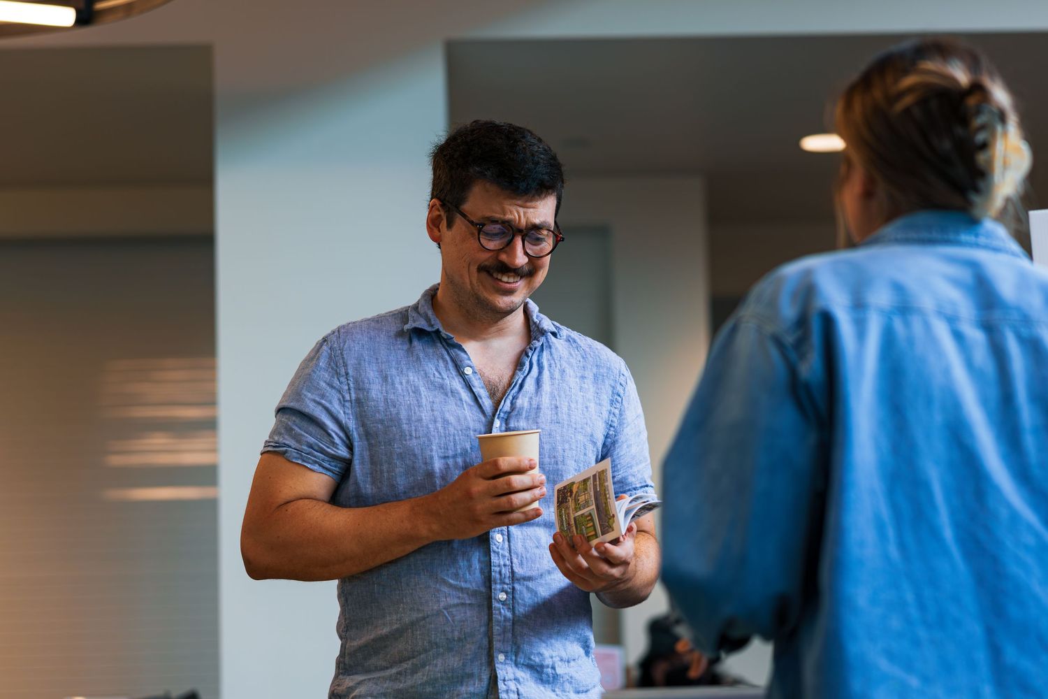 Person in a blue button-down shirt holds a mobile phone while standing in an office hallway.