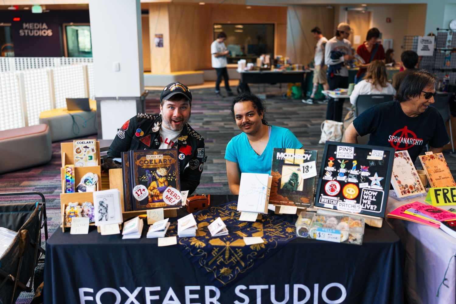 Artists display merchandise and artwork at their booth during a convention with a Foxfaer Studio banner.