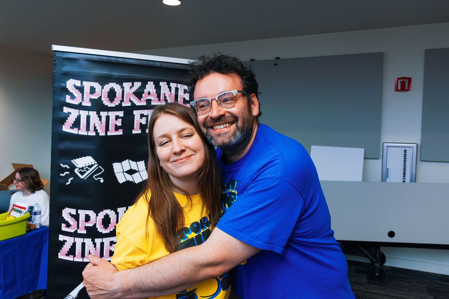 Two people embrace and smile at a Spokane Zine event with a black banner visible in the background.