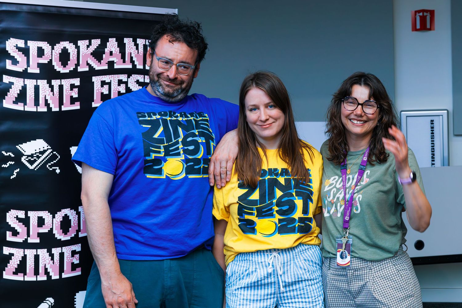 People gather at a zine fest event with blue t-shirts and promotional signage visible in the background.