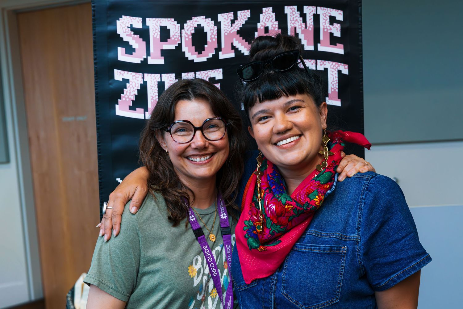 Two people smiling together in front of a Spokane sign wearing casual attire and colorful accessories.