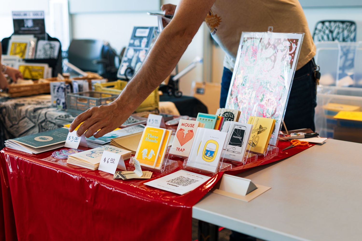 Hands browse through colorful greeting cards and stationery displayed on a red tablecloth at a craft market.