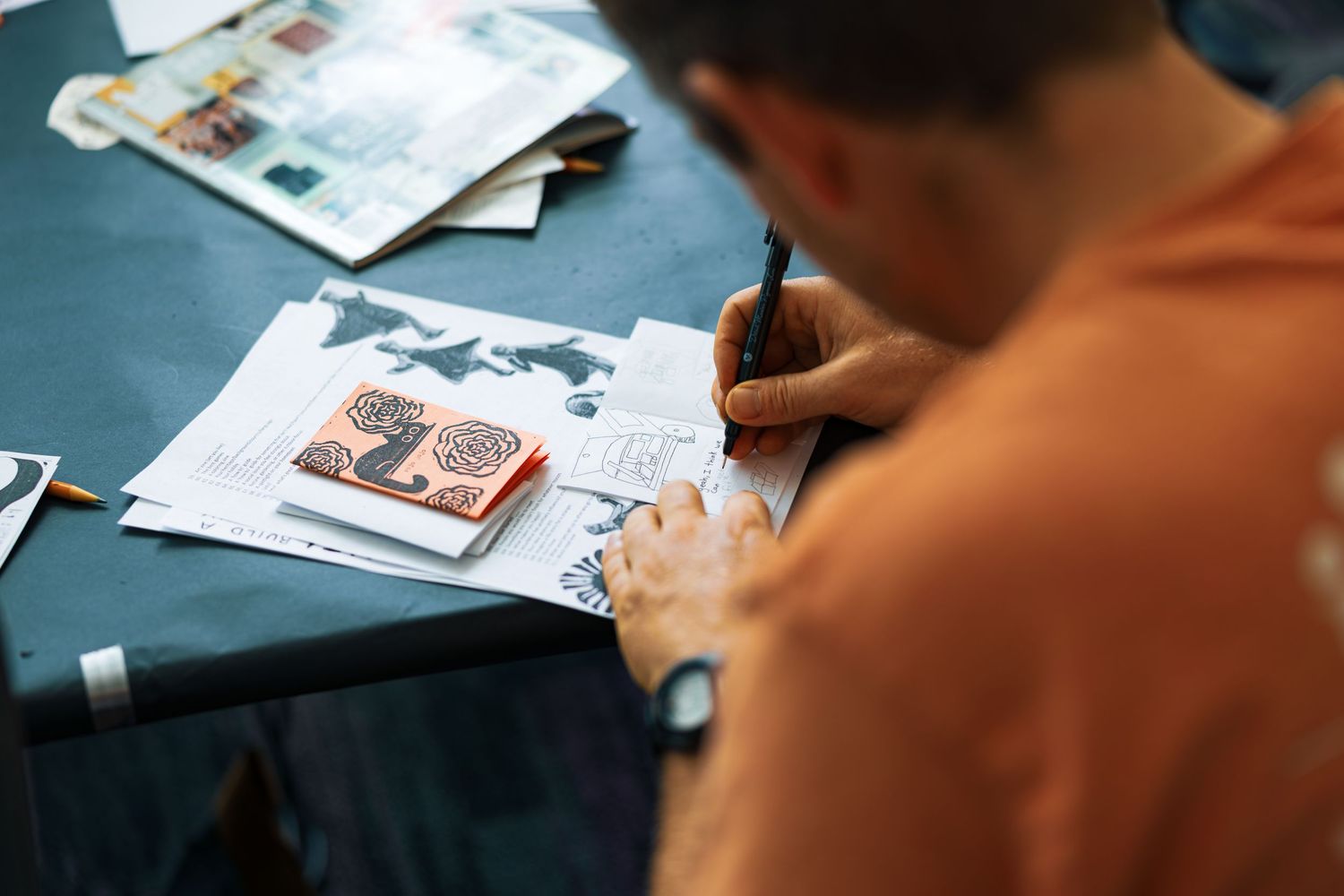 Someone writes in a notebook with a pen while papers and documents are spread across a blue desk surface.