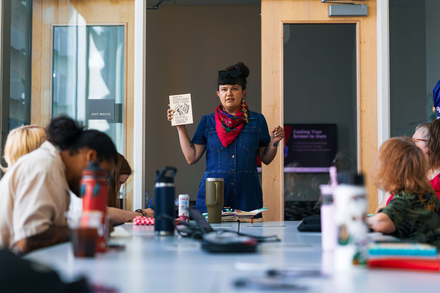 A person wearing a bandana and denim shirt stands at the head of a table during what appears to be a meeting or workshop.