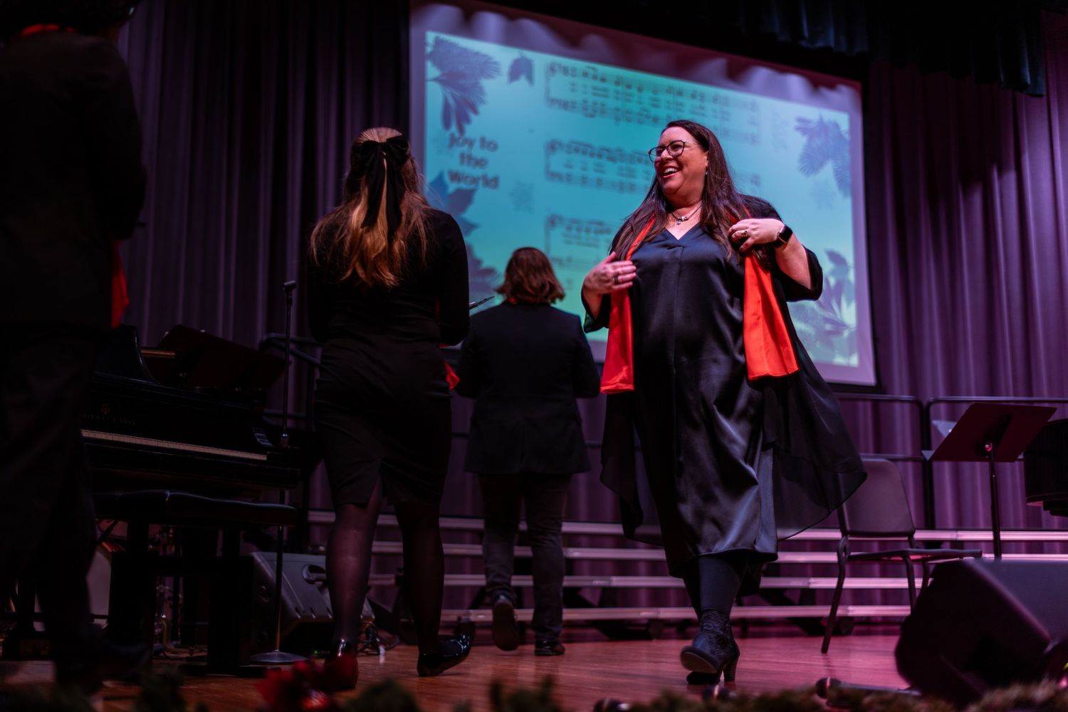 A performer in dark clothing sings on stage with a projection screen and other performers visible in the background.
